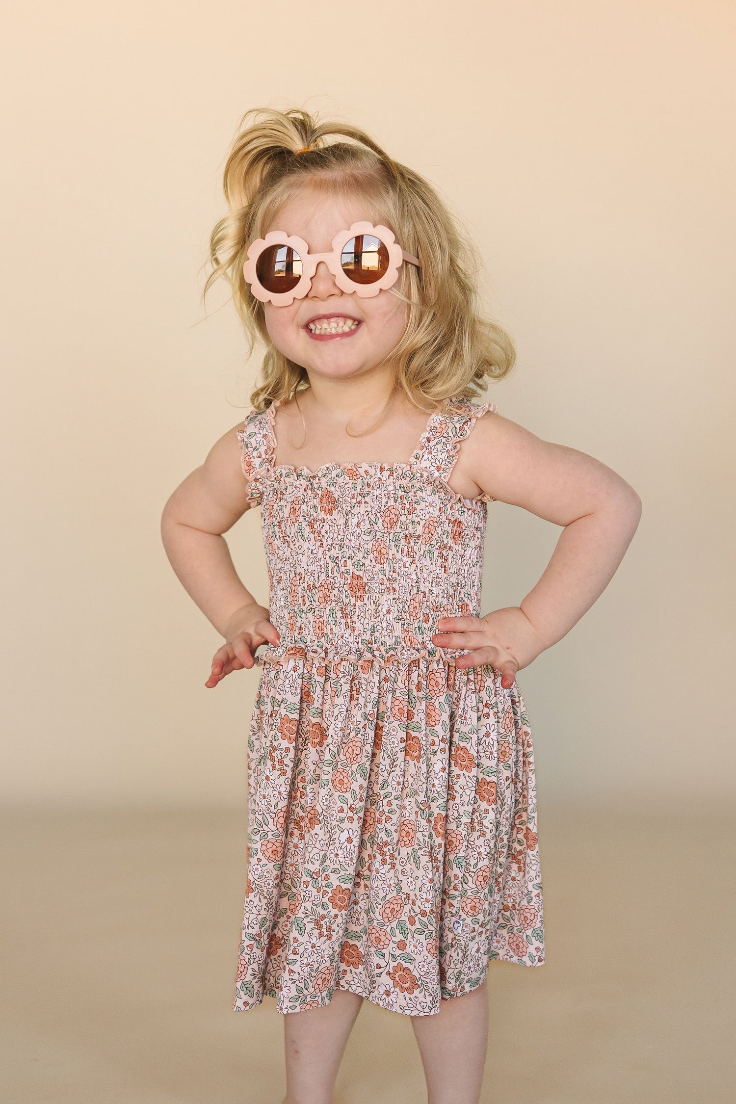 Smiling toddler wearing a floral smocked sundress by little nessi and pink flower-shaped sunglasses, standing with hands on hips against a neutral background.